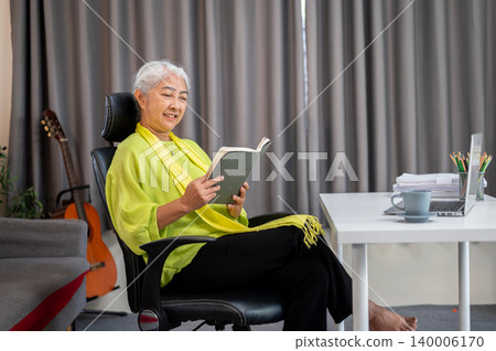 Happy elderly old asian woman reading a book sitting crossed legs on chair in a living room at home. 140006170