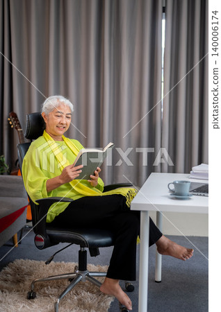 Full body of elderly old asian woman reading a book on chair with bare foots in living room at home. Full body of elderly old asian woman reading a book on chair with bare foots in living room at home. 140006174