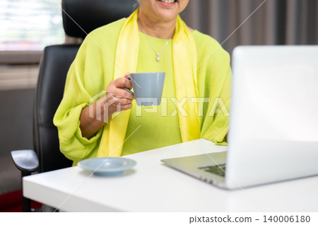 Close up of smiling elderly old asian woman holding coffee looking at laptop on table in home office Close up of smiling elderly old asian woman holding coffee looking at laptop on table in home office 140006180