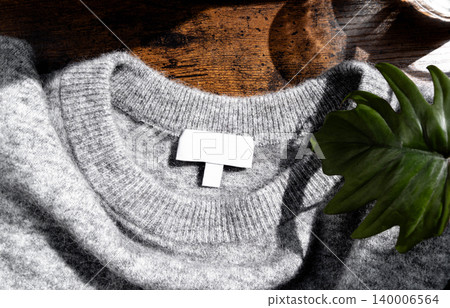 Gray knitted sweater with blank clothing label on a wooden surface and green leaf Gray knitted sweater with blank clothing label on a wooden surface and green leaf 140006564