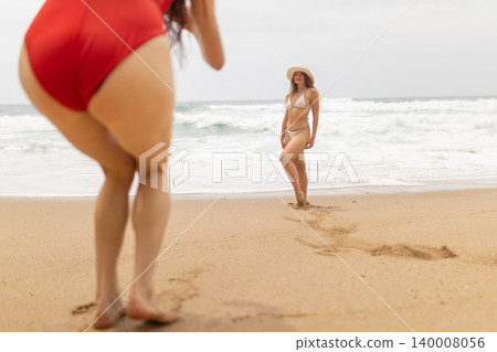 Two friends enjoy a day at the beach taking pictures and having fun in the sand near the ocean on a cloudy day 140008056
