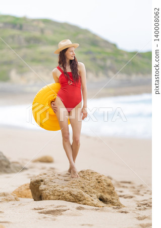 Woman stands on rock with a yellow float at beach, enjoying the ocean view and summer sky Woman stands on rock with a yellow float at beach, enjoying the ocean view and summer sky 140008062