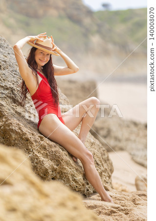 Woman in red swimsuit enjoys time on rocky beach while wearing straw hat during sunny day Woman in red swimsuit enjoys time on rocky beach while wearing straw hat during sunny day 140008070