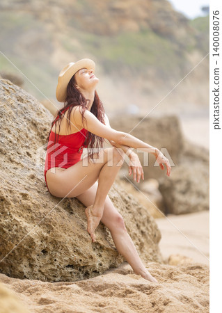 Woman sits on a rock at the beach during a sunny day and enjoys the warm weather while wearing a swimsuit and hat Woman sits on a rock at the beach during a sunny day and enjoys the warm weather while wearing a swimsuit and hat 140008076