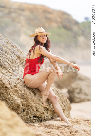 Woman sitting on a rock at the beach wearing a red swimsuit and a hat enjoying the warm weather during the day Woman sitting on a rock at the beach wearing a red swimsuit and a hat enjoying the warm weather during the day 140008077
