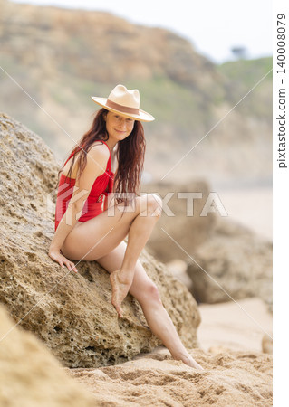 Woman in red swimsuit sits on rocks at beach during daytime with ocean in background and tan sand 140008079