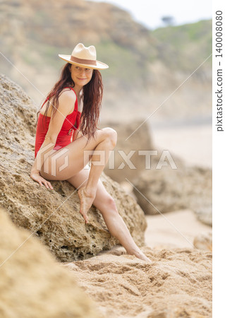Woman sits on a rock by the beach wearing a red swimsuit and hat during a sunny day in a coastal area Woman sits on a rock by the beach wearing a red swimsuit and hat during a sunny day in a coastal area 140008080