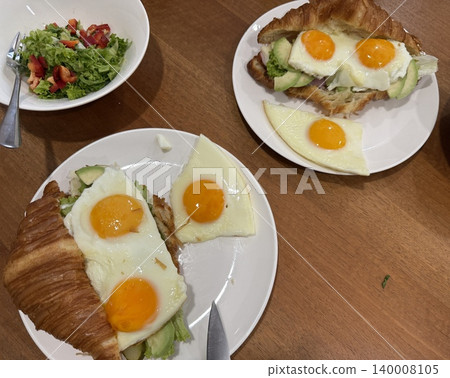 Breakfast croissant sandwich with fried eggs and avocado on white plates with fresh salad added. Breakfast croissant sandwich with fried eggs and avocado on white plates with fresh salad added. 140008105