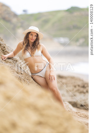 Woman in white bikini poses on sandy beach near rocky cliffs during sunny day Woman in white bikini poses on sandy beach near rocky cliffs during sunny day 140008129