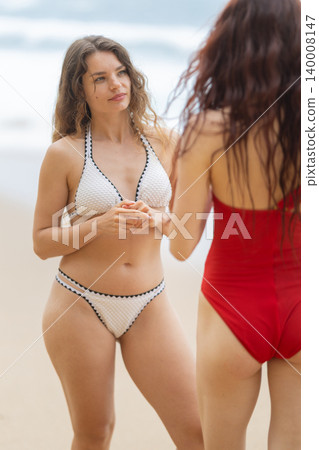 Two women chat and enjoy the beach on a sunny day while wearing swimwear and standing on the sand near the ocean 140008147