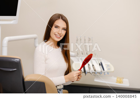 Pretty girl smiling while sitting in the dentist's chair, holding a mirror in her hands Pretty girl smiling while sitting in the dentist's chair, holding a mirror in her hands 140008845
