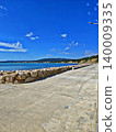 A scenic seaside landscape featuring a calm blue sea, a protective stone breakwater extending into the water, and a clear summer sky, evoking a sense of tranquility and coastal beauty. 140009335