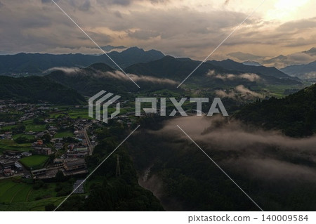 An aerial photograph overlooking a mountain village shrouded in mist and an arch bridge in Hinokage Town, Kumamoto Prefecture. An aerial photograph overlooking a mountain village shrouded in mist and an arch bridge in Hinokage Town, Kumamoto Prefecture. 140009584