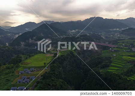 An aerial photograph overlooking a misty mountain village and a red bridge in Hinokage Town, Kumamoto Prefecture. An aerial photograph overlooking a misty mountain village and a red bridge in Hinokage Town, Kumamoto Prefecture. 140009703