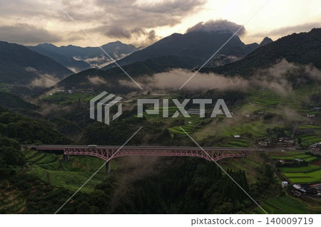 一張空拍照片,展現了宮崎縣日之影町一座橫跨霧氣繚繞的山谷的紅色拱橋。 一張空拍照片,展現了宮崎縣日之影町一座橫跨霧氣繚繞的山谷的紅色拱橋。 140009719