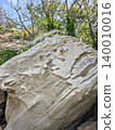 A large, weathered sandstone boulder with intricate natural texture, nestled at the edge of a forest with sun-drenched trees in the background, showcasing a serene outdoor landscape. 140010016