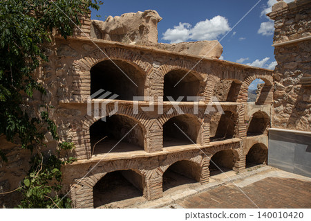A row of empty arched brick crypts and stone burial niches is visible in a historic Spanish cemetery. The sky above is blue with some clouds. A row of empty arched brick crypts and stone burial niches is visible in a historic Spanish cemetery. The sky above is blue with some clouds. 140010420