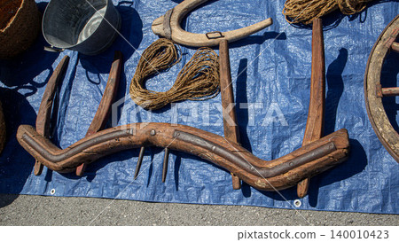 Traditional vintage wooden ox yokes and coils of rope are arranged on a blue tarp at a rustic flea market. Many people browse and explore various items for sale. Traditional vintage wooden ox yokes and coils of rope are arranged on a blue tarp at a rustic flea market. Many people browse and explore various items for sale. 140010423