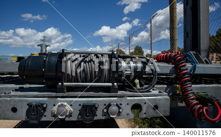 An electric towing winch is mounted on an empty transport trailer. The trailer is placed outdoors under a clear blue sky with white clouds in the background. An electric towing winch is mounted on an empty transport trailer. The trailer is placed outdoors under a clear blue sky with white clouds in the background. 140011576