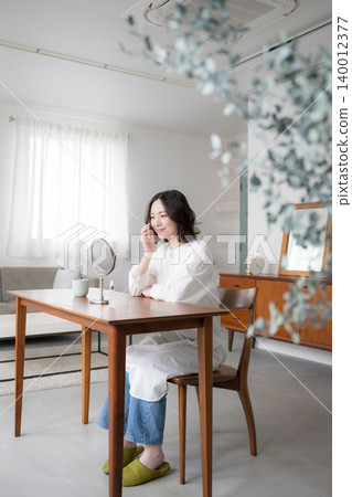 A woman enjoying a mindful lifestyle in her living room, smiling as she looks in the mirror while doing her skincare routine, and her focus on beauty and a natural lifestyle. A woman enjoying a mindful lifestyle in her living room, smiling as she looks in the mirror while doing her skincare routine, and her focus on beauty and a natural lifestyle. 140012377