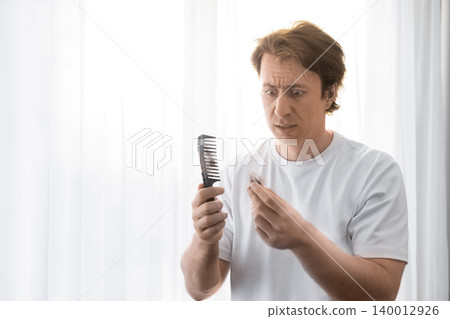 A stressed man looks in shock at a comb and fallen hair. This image highlights male hair loss, alopecia, and the testosterone concept during his daily grooming routine. A stressed man looks in shock at a comb and fallen hair. This image highlights male hair loss, alopecia, and the testosterone concept during his daily grooming routine. 140012926