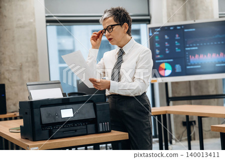 Businesswoman reviewing printed documents at a modern office workspace, with a printer and digital display showcasing data analysis and charts Businesswoman reviewing printed documents at a modern office workspace, with a printer and digital display showcasing data analysis and charts 140013411