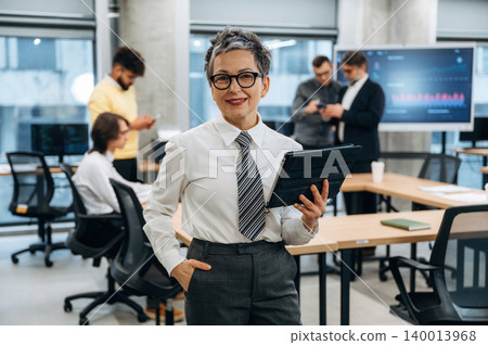 Confident businesswoman holding tablet in modern office, surrounded by colleagues engaged in discussions, showcasing teamwork and collaboration in a professional environment Confident businesswoman holding tablet in modern office, surrounded by colleagues engaged in discussions, showcasing teamwork and collaboration in a professional environment 140013968