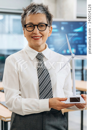 Businesswoman in formal attire holding a miniature wind turbine model in a modern office, showcasing commitment to sustainability and renewable energy solutions Businesswoman in formal attire holding a miniature wind turbine model in a modern office, showcasing commitment to sustainability and renewable energy solutions 140014126
