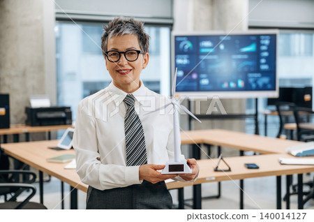 Businesswoman proudly holding a miniature wind turbine model in a modern office, showcasing commitment to sustainability and renewable energy solutions Businesswoman proudly holding a miniature wind turbine model in a modern office, showcasing commitment to sustainability and renewable energy solutions 140014127