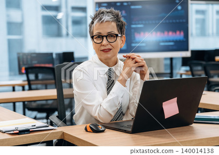 Confident businesswoman in a modern office setting, seated at a desk with a laptop, showcasing professionalism and engagement in her work environment 140014138