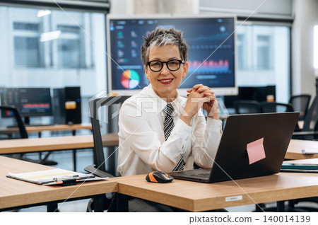 Businesswoman in modern office setting, smiling confidently at her laptop, showcasing professional attire and a vibrant workspace with data charts in the background 140014139