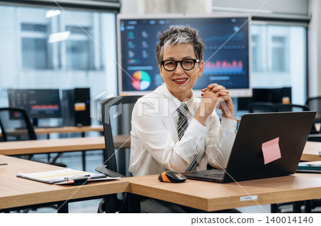Businesswoman in modern office setting, smiling while working on laptop, showcasing professional attire and engaging workspace with data visuals in background 140014140