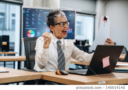 Excited businesswoman celebrating success at her desk in a modern office, holding documents and showing enthusiasm for her achievements in a professional environment 140014148