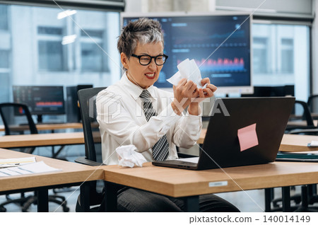 Businesswoman in office expressing frustration while working on laptop, surrounded by modern workspace elements, illustrating the challenges of corporate life 140014149