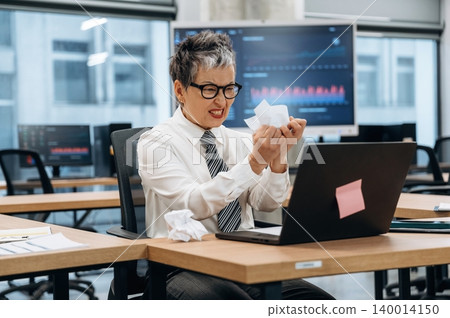 Businesswoman in modern office expressing frustration while working on laptop, surrounded by data charts and crumpled papers, illustrating workplace stress and challenges Businesswoman in modern office expressing frustration while working on laptop, surrounded by data charts and crumpled papers, illustrating workplace stress and challenges 140014150