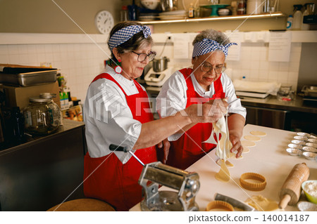 Senior Caucasian and Black women preparing fresh pasta dough in a commercial kitchen setting Senior Caucasian and Black women preparing fresh pasta dough in a commercial kitchen setting 140014187