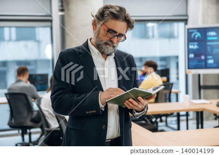 Mature businessman with glasses is reviewing notes in a modern office environment, showcasing collaboration and productivity among colleagues in the background Mature businessman with glasses is reviewing notes in a modern office environment, showcasing collaboration and productivity among colleagues in the background 140014199