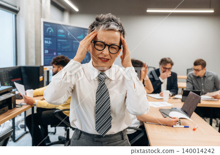 Businesswoman in a modern office is experiencing stress while working, surrounded by colleagues engaged in discussions and using technology, illustrating workplace pressure Businesswoman in a modern office is experiencing stress while working, surrounded by colleagues engaged in discussions and using technology, illustrating workplace pressure 140014242