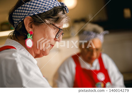 Senior Caucasian woman focuses on food preparation in a bright modern kitchen with a senior colleague Senior Caucasian woman focuses on food preparation in a bright modern kitchen with a senior colleague 140014279