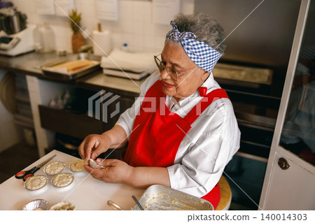 Senior African American woman preparing handmade pastries in a professional commercial kitchen bakery. 140014303
