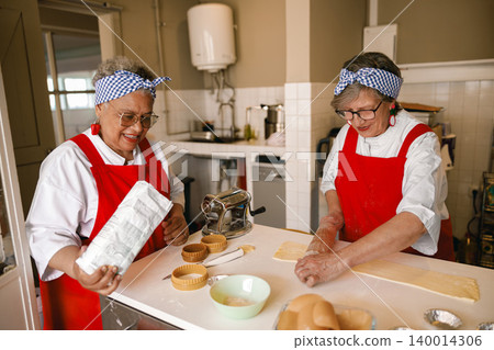 Diverse senior Black and Caucasian women preparing dough in a bright professional kitchen setting Diverse senior Black and Caucasian women preparing dough in a bright professional kitchen setting 140014306