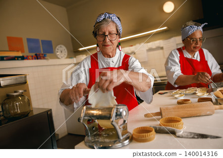 Senior Caucasian and African American women rolling fresh dough in a professional kitchen environment Senior Caucasian and African American women rolling fresh dough in a professional kitchen environment 140014316