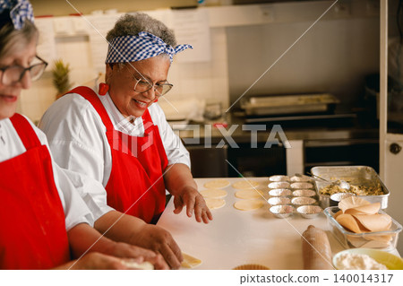 Diverse senior Black and White women in red aprons baking pastries inside a bright professional kitchen Diverse senior Black and White women in red aprons baking pastries inside a bright professional kitchen 140014317