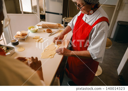 Senior Caucasian woman cutting circles of fresh pasta dough in a bright professional kitchen setting Senior Caucasian woman cutting circles of fresh pasta dough in a bright professional kitchen setting 140014323