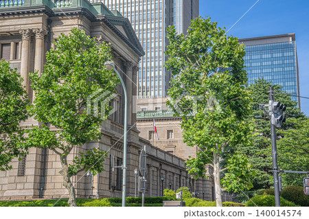 The exterior of the Bank of Japan headquarters, the lush green street trees, and the modern skyscrapers in the background [Nihonbashi Honcho, Chuo-ku, Tokyo] 140014574
