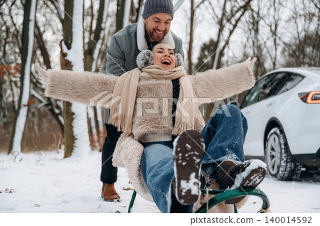 Joyful couple enjoying winter fun, with one person sledding down a snowy hill while the other pushes, capturing the essence of outdoor happiness and playfulness 140014592