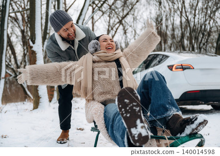 Joyful couple enjoying winter fun while sledding in snowy landscape, showcasing laughter and excitement in a playful outdoor setting with trees 140014598
