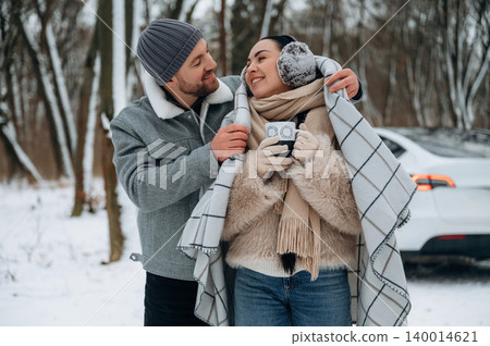 Couple enjoying warm beverages while wrapped in a cozy blanket outdoors in winter, surrounded by snow-covered trees, capturing a moment of love and warmth Couple enjoying warm beverages while wrapped in a cozy blanket outdoors in winter, surrounded by snow-covered trees, capturing a moment of love and warmth 140014621