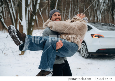 Happy couple enjoying winter day outdoors, man playfully lifting woman in snowy forest, showcasing joy and connection in a beautiful winter landscape 140014626