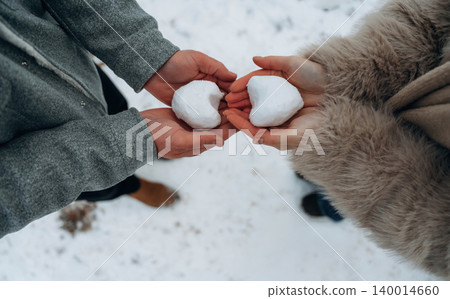 Two individuals holding snowballs shaped like hearts, showcasing a playful winter moment in a snowy outdoor setting, evoking warmth and connection 140014660